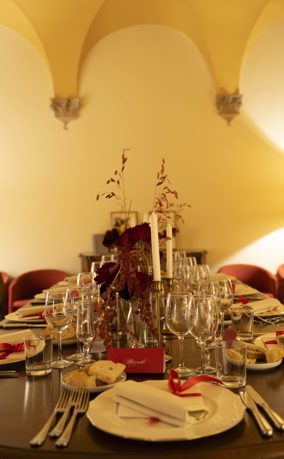 Tavola apparecchiata con fiori bordeaux, candele e segnaposto sotto volta a crociera